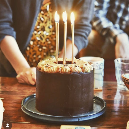 A chocolate birthday cake with 6 light candles sits upon a table surrounded by a group of people