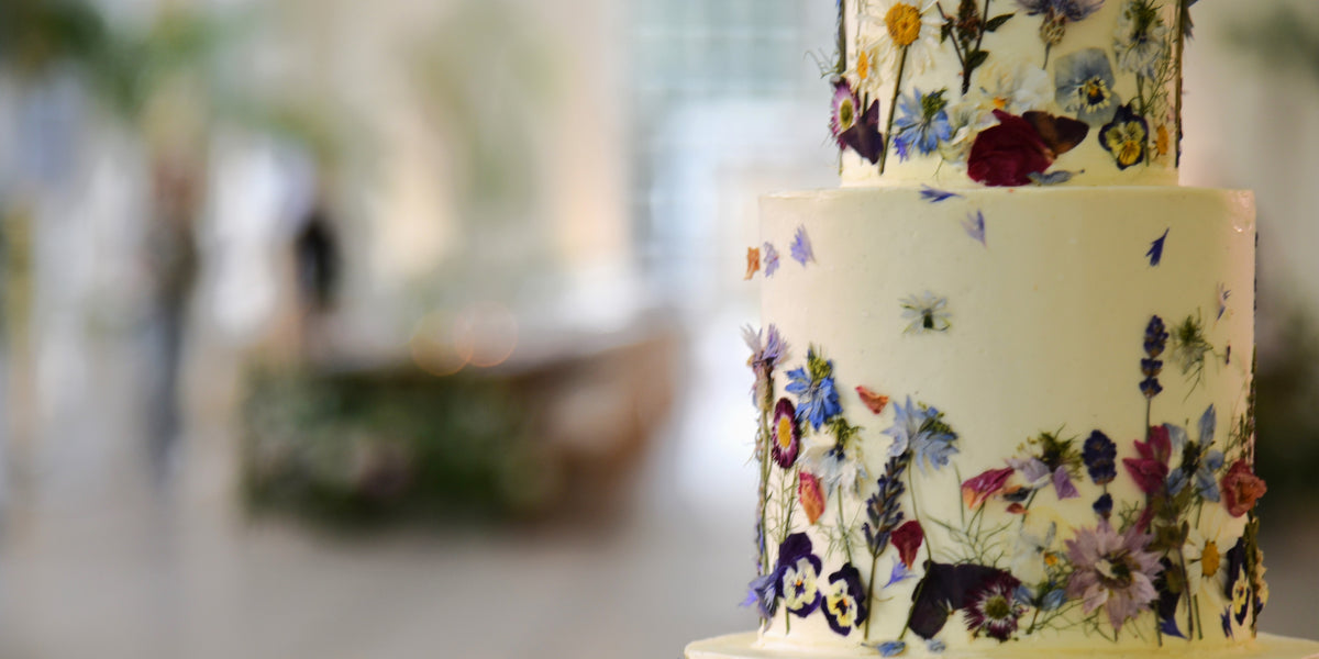 A two tier cake decorated with blue flowers sits at the forefront of a blurred background